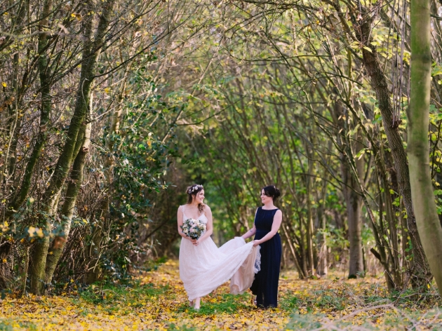 Bride in a white dress and a bridesmaid in black dress walk through the woods