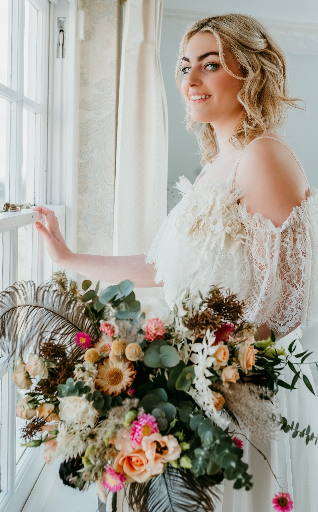 Boho bride stood by a window with loose, bohemian bouquet