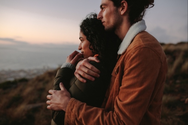 couple in coats on a hillside looking out at the view the same way he is hugging her 