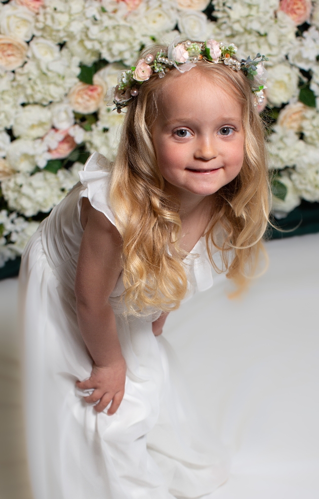 flowergirl in white dress in front of flowerwall