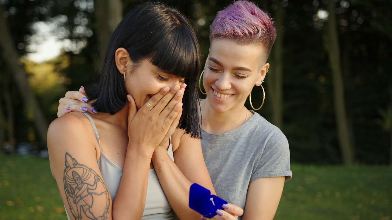 two women who have just got engaged one standing with her hands to her mouth, the other holding the ring