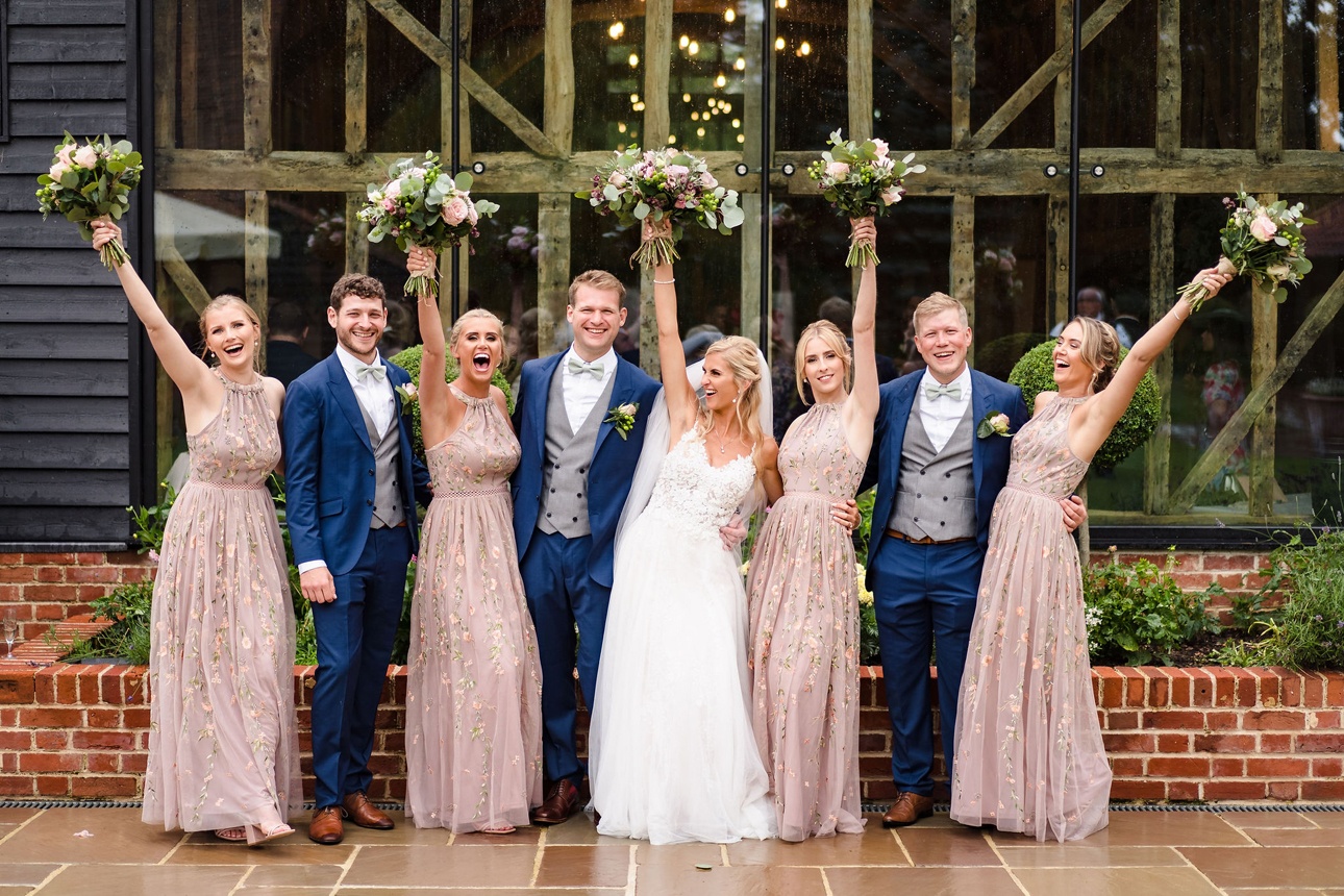 Whole bridal party stands outside venue with bouquets held aloft