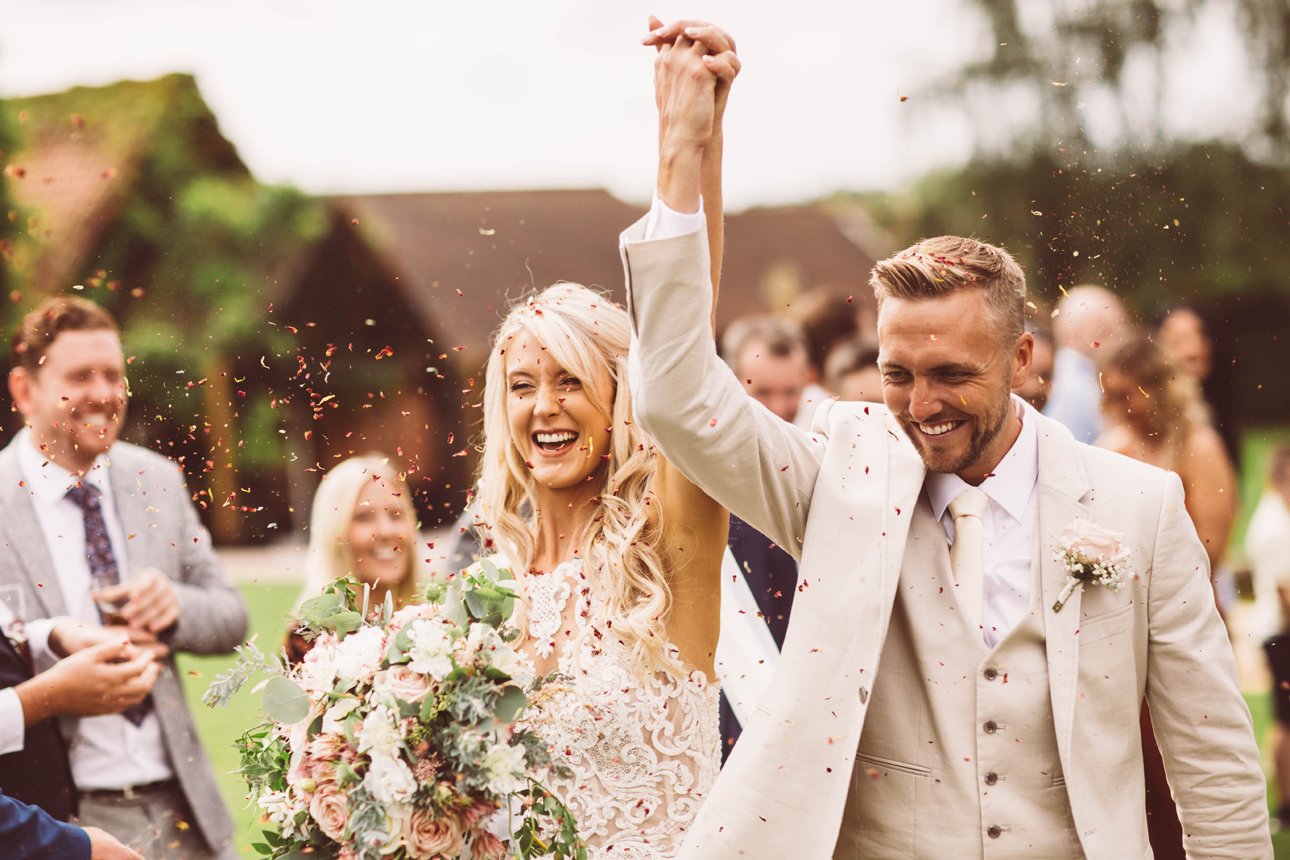 Bride and groom hold hands aloft as they walk through confetti