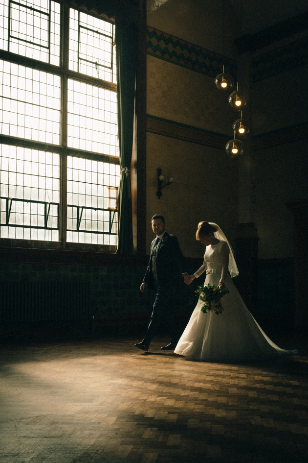 Bride and groom walk through dark hall towards bright window
