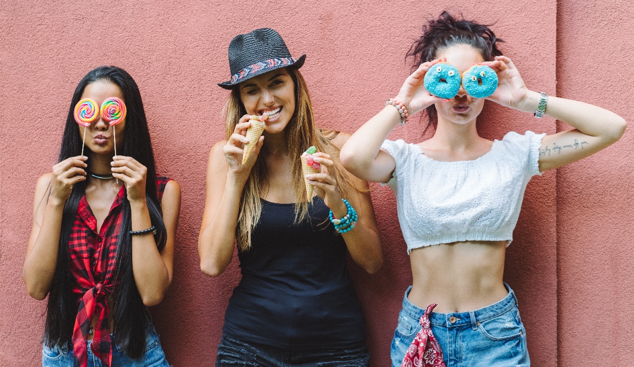 three people standing in front of a pink wall one with lollipops one with ice cream and one with doughnuts 