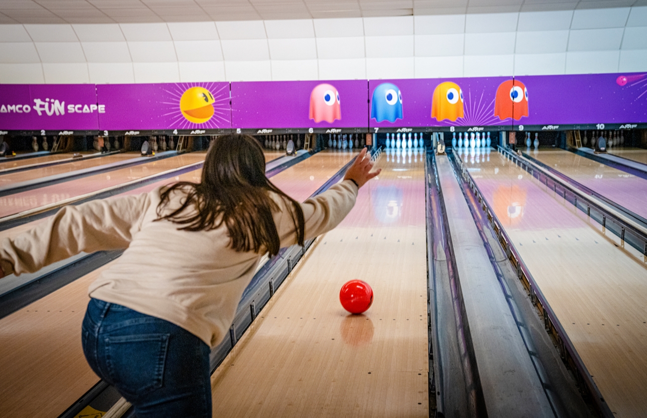 woman in jeans and cream jumper throwing a bowling ball down the alley towards pins