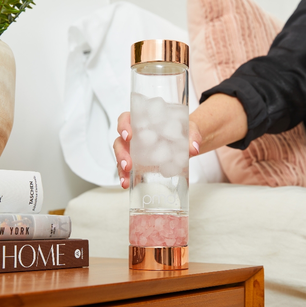 clear water bottle with rose gold top and bottom with rose quartz in the base, resting next to bed on cabinet