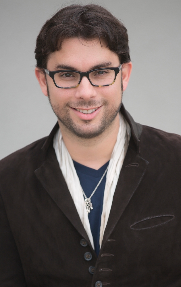 Roberto Valenzuela in a brown jacket shirt and tshirt wearing a necklace and reading glasses