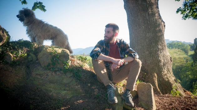 Young man with a beard and woodcutter shirt sitting in the countryside with his Catalan shepherd dog