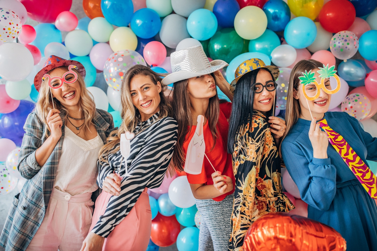five women pose with photo booth props in front of a balloon wall 