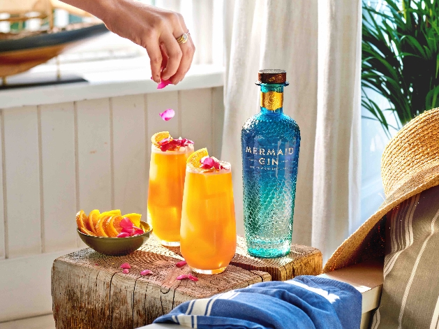bottle of blue gin with two cocktails next to it of orange juice hand sprinkling in petals