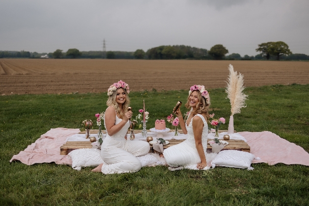 Two females sat on a white blanket sipping champagne