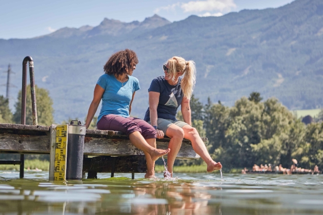 two women sat at the end of a pier soaking their feet in water 