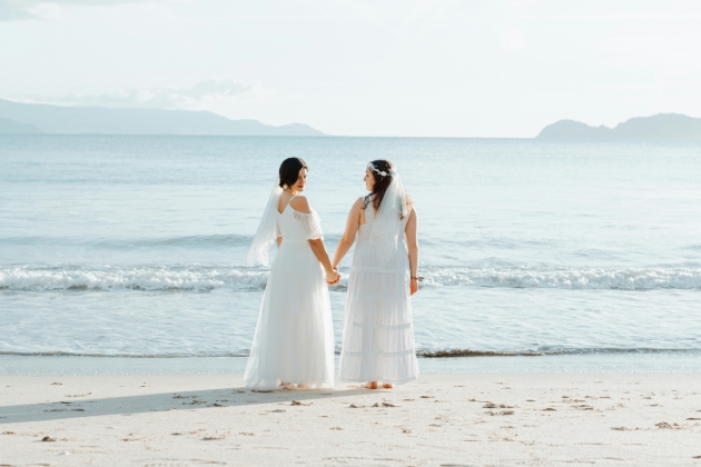 two women in wedding dresses on the beach holding hands